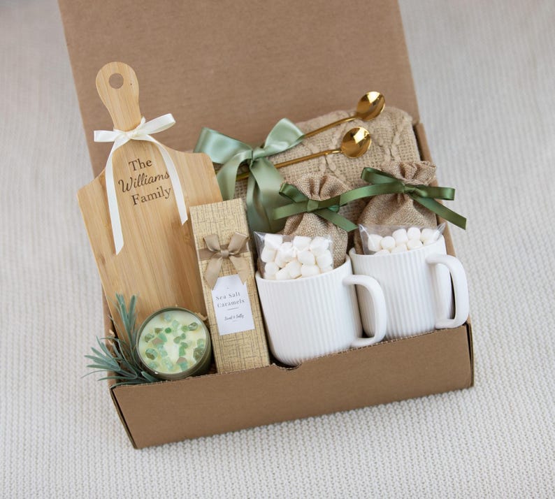 May include: A gift box containing a wooden paddle board with the text The Williams Family, a candle with green stones, a box of Sea Salt Caramels, two white ribbed mugs filled with marshmallows in burlap bags, and two gold spoons with a green ribbon.