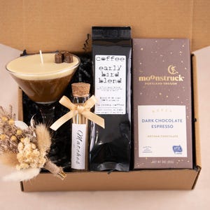 May include: A gift box containing a coffee-themed assortment. Includes a candle in a martini glass, a bag of coffee, a dark chocolate bar, a vial of matches, and a small dried flower arrangement. The coffee bag reads "early bird blend".