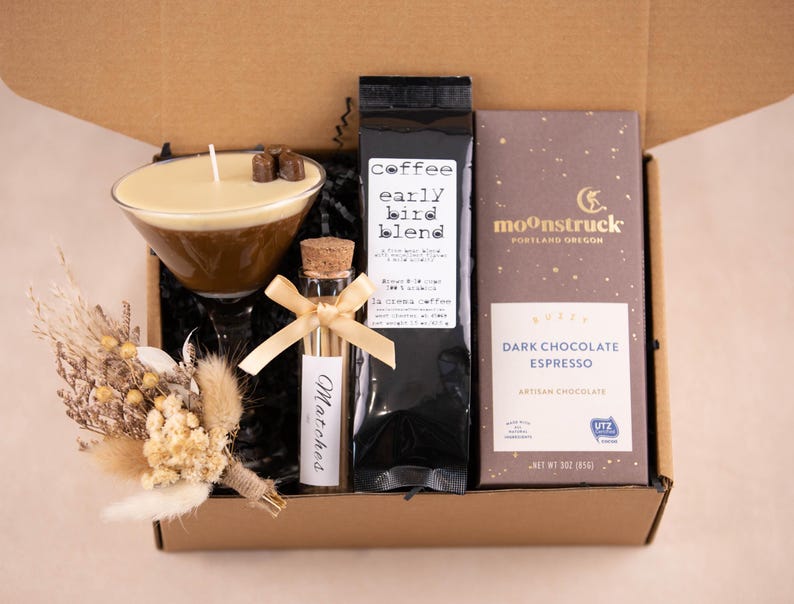 May include: A gift box containing a coffee-themed assortment. Includes a candle in a martini glass, a bag of coffee, a dark chocolate bar, a vial of matches, and a small dried flower arrangement. The coffee bag reads "early bird blend".