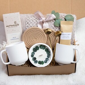 Puede incluir: Caja de regalo llena de una taza blanca, barra de chocolate, caf&eacute;, vela, posavasos, cucharas doradas y una manta de punto atada con una cinta. La caja es marr&oacute;n y est&aacute; sobre una superficie blanca y esponjosa.