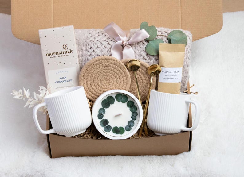 Puede incluir: Caja de regalo llena de una taza blanca, barra de chocolate, caf&eacute;, vela, posavasos, cucharas doradas y una manta de punto atada con una cinta. La caja es marr&oacute;n y est&aacute; sobre una superficie blanca y esponjosa.
