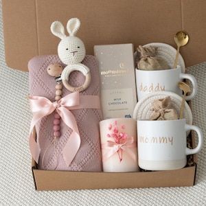 May include: A gift box filled with baby items. Includes a pink blanket, a crochet bunny, a wooden teether, a chocolate bar, a candle, and two mugs that say "daddy" and "mommy". The box also contains two gold spoons.