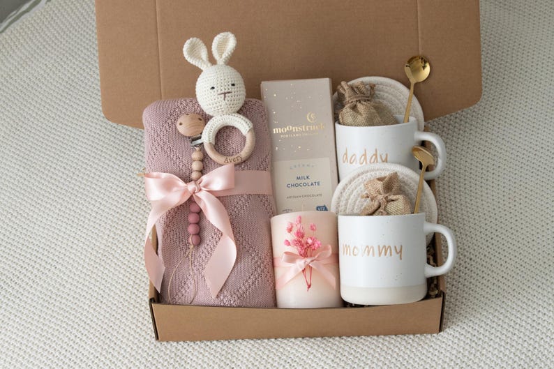 May include: A gift box filled with baby items. Includes a pink blanket, a crochet bunny, a wooden teether, a chocolate bar, a candle, and two mugs that say "daddy" and "mommy". The box also contains two gold spoons.