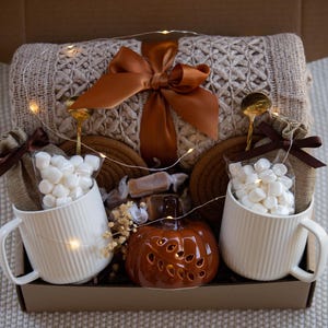 Puede incluir: Una caja de regalo llena con una manta de punto beige, un lazo marr&oacute;n grande, dos tazas blancas con malvaviscos, una calabaza de cer&aacute;mica y cucharas doradas. La caja tambi&eacute;n contiene caramelos y luces decorativas.