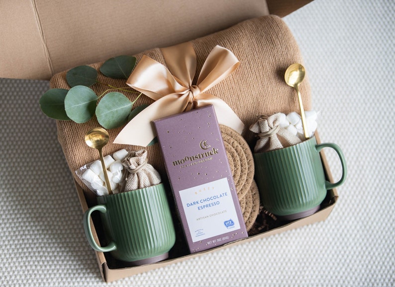 May include: A gift box with a tan blanket, a gold ribbon, and eucalyptus sprigs. It contains two green mugs, gold spoons, marshmallows, and a bar of dark chocolate espresso. The chocolate bar reads "Moonstruck" and "Dark Chocolate Espresso".