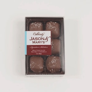 May include: A clear plastic box containing five milk chocolate sea salt caramels. The caramels are square-shaped and sprinkled with sea salt. The box has a label that reads "Jason & Mary's" and "Milk Chocolate Sea Salt Caramels, 3 oz (85g)."