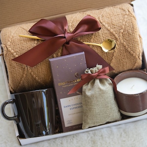 May include: A gift box filled with cosy items for relaxation, including a brown knitted throw blanket, a brown mug, a bar of dark chocolate espresso, a small hessian bag with a brown ribbon, and a brown and white candle.
