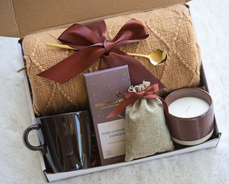 May include: A gift box filled with cosy items for relaxation, including a brown knitted throw blanket, a brown mug, a bar of dark chocolate espresso, a small hessian bag with a brown ribbon, and a brown and white candle.