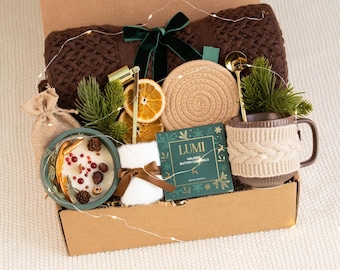 May include: A gift box containing a dark brown knit blanket with a green velvet bow, a candle in a green bowl, a beige rope coaster, a mug with a knit sleeve, a gold spoon, a box of Lumi Holiday Butter Caramels, and a small burlap pouch.