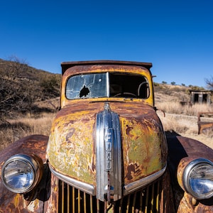 May include: A rusty yellow vintage Ford truck with a broken windshield and a chrome grill. The truck is parked in a desert landscape.