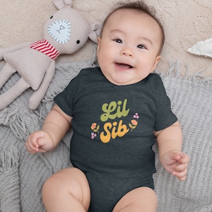 Op de afbeelding: Een lachende baby draagt een donkergrijze romper met de tekst "Lil Sib" in geel en oranje, versierd met kleine bloemen. De baby ligt op een grijze deken met een knuffel en een wit stervormig kussen.