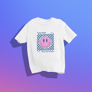 May include: White t-shirt featuring a pink smiley face within a blue and white checkered square. The text "THEY/THEM" and "OR JUST SAY MY NAME!" are printed above and below the smiley face design.