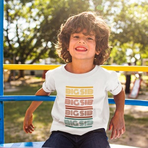 Pode incluir: Uma camiseta branca com o texto "BIG SIB" em um design retrô empilhado. O texto está em uma variedade de cores, incluindo amarelo, laranja, vermelho e verde. A camiseta é usada por uma criança com cabelo castanho encaracolado.