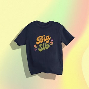 May include: Navy blue t-shirt with the words "Big Sib" in orange and green retro font, decorated with small floral accents. The shirt is a classic crew neck style.