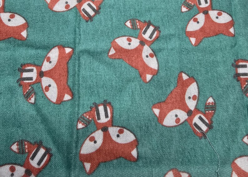 May include: Teal fabric with a repeating pattern of cartoon foxes in red, white, and black. The foxes have white faces, red bodies, and black and white striped tails. The fabric has a textured appearance.
