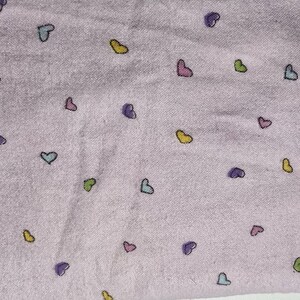 May include: A light purple fabric with a pattern of small, colorful hearts. The hearts are outlined in black and filled with shades of yellow, green, blue, pink, and purple. The fabric has a soft, slightly textured appearance.