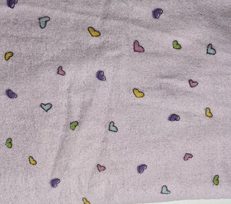 May include: A light purple fabric with a pattern of small, colorful hearts. The hearts are outlined in black and filled with shades of yellow, green, blue, pink, and purple. The fabric has a soft, slightly textured appearance.