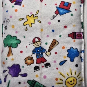 May include: A white rectangular fabric item with a colorful cartoon design. The design includes a baseball player, crayons, paint splatters, a tree, a sun, and a kite. The item is approximately 3 inches wide.