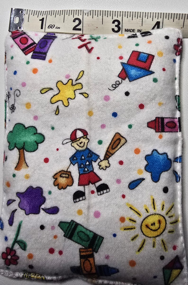 May include: A white rectangular fabric item with a colorful cartoon design. The design includes a baseball player, crayons, paint splatters, a tree, a sun, and a kite. The item is approximately 3 inches wide.