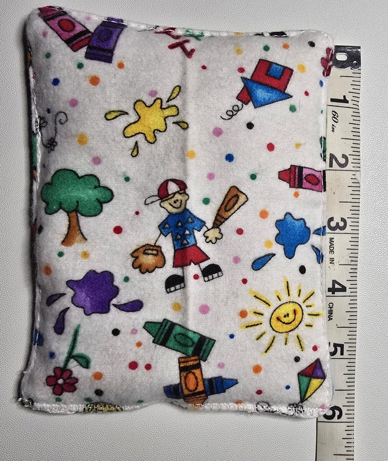 May include: A white rectangular pillow with a colorful print of crayons, baseball, a tree, and a smiling sun. The pillow is approximately 5 inches long.