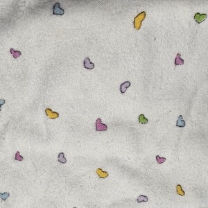 May include: White fabric with a pattern of small, colorful hearts in shades of blue, pink, yellow, and green. The hearts are scattered across the surface, creating a playful and whimsical design. The fabric appears to be soft and textured.