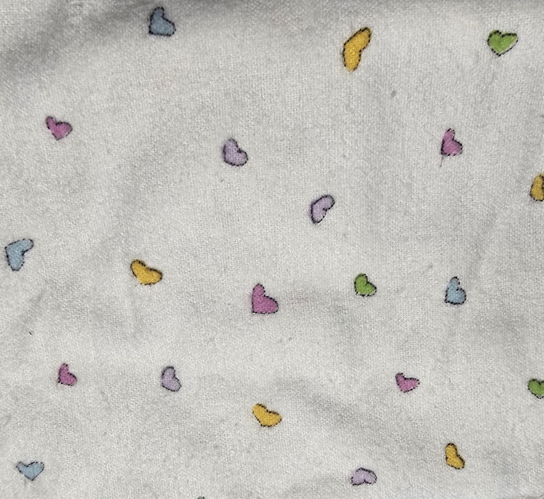 May include: White fabric with a pattern of small, colorful hearts in shades of blue, pink, yellow, and green. The hearts are scattered across the surface, creating a playful and whimsical design. The fabric appears to be soft and textured.
