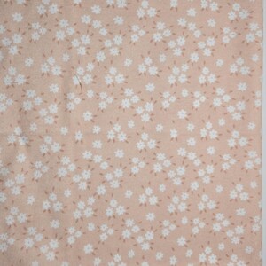 May include: A close-up of a fabric with a repeating floral pattern. The fabric has a light peach background with small white flowers and tiny brown accents. The flowers are clustered together, creating a delicate and feminine design.