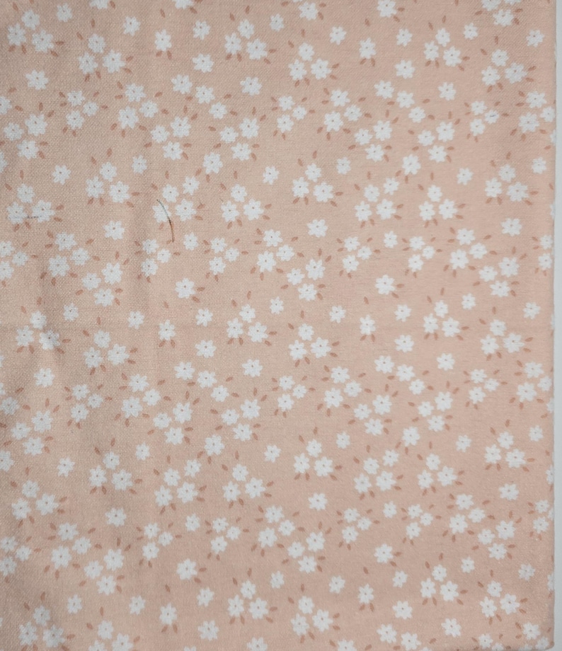 May include: A close-up of a fabric with a repeating floral pattern. The fabric has a light peach background with small white flowers and tiny brown accents. The flowers are clustered together, creating a delicate and feminine design.
