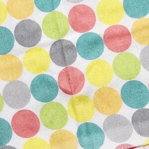 May include: Close-up of a white fabric with a repeating pattern of large, colorful polka dots. The dots are in shades of yellow, pink, green, blue, and gray. The fabric appears to be soft and textured.