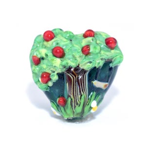 May include: A handcrafted glass bead shaped like a miniature apple tree. The tree features a green canopy with red apples, a brown trunk, and a dark blue base with green grass and small white flowers. The bead is designed to be a decorative element.