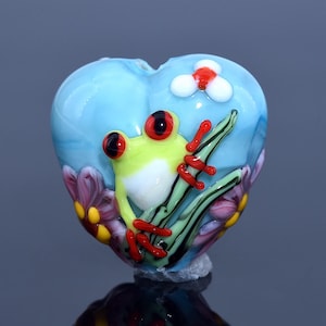 Peut inclure: Sculpture en verre en forme de cœur représentant une grenouille verte aux yeux et pieds rouges, posée sur des feuilles vertes et noires. La grenouille est sur un fond bleu clair avec des fleurs blanches et rouges et des fleurs violettes.