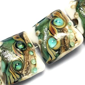 May include: Close-up of a set of four square-shaped glass beads with a white, green, and brown marbled pattern. The beads have a glossy finish and feature small, turquoise-colored accents.