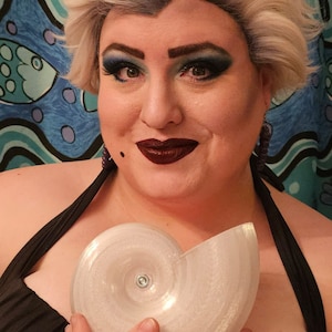 May include: A person dressed as Ursula from The Little Mermaid, wearing a black swimsuit and holding a white seashell prop.