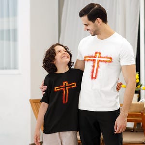 Puede incluir: Una camiseta blanca de manga corta con un gráfico de cruz naranja y roja brillante en el pecho, combinada con una camiseta negra de manga corta con un gráfico similar de cruz naranja y roja brillante en el pecho, hechas de algodón.