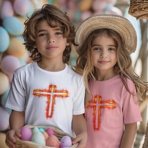 Puede incluir: Una camiseta blanca y una camiseta rosa, cada una con un gráfico de una cruz delineada con llamas naranjas y amarillas. La camiseta blanca la lleva un niño de pelo castaño ondulado y la rosa un niño de pelo largo y rubio.