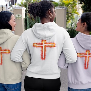 Puede incluir: Tres sudaderas con capucha en beige, blanco y gris claro, cada una con un estampado gráfico en la espalda de una cruz blanca delineada por llamas de color naranja y rojo vibrantes. Las sudaderas tienen mangas largas y capucha.