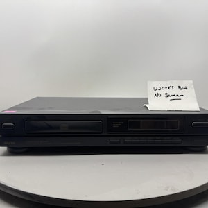 May include: A black Sanyo CD player with a front-loading disc tray, a digital display panel, and several control buttons. A white note is placed on top of the device with the handwritten text "Works But No Screen" in black ink.