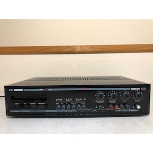 May include: Black CMDS compact system audio receiver with multiple knobs and buttons. The receiver has a series of dials for microphone volume, program volume, bass, and treble. The front panel also features a series of buttons and a slot for a CD or tape.