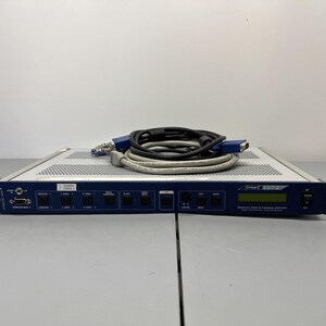 May include: A blue and silver rack-mount video switcher with a front control panel featuring multiple buttons, a small digital display, and a power switch. A coiled black and gray VGA cable with blue connectors rests on top of the metal chassis.