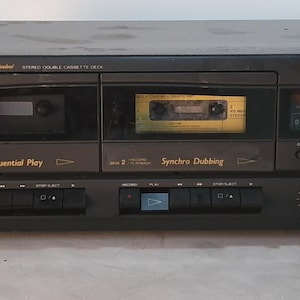 May include: A black Fisher Studio-Standard stereo double cassette deck featuring two tape compartments, one containing a yellow cassette tape. The front panel displays text including Sequential Play, Synchro Dubbing, and model number PH-W60J, with various control buttons and knobs.