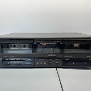 May include: A black Technics stereo double cassette deck. The deck has two cassette slots, a display screen, and various control buttons. The cassette deck is on a white surface.