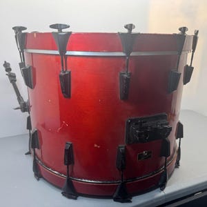 May include: A cylindrical red drum with a glossy finish, featuring black metal hardware, tension lugs, and a mounting bracket. The drum shell has a visible metallic red hue with some minor surface wear and a small rectangular black badge.