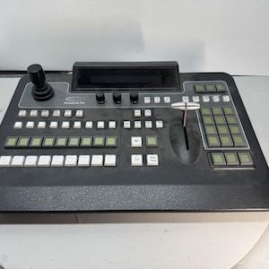 May include: A black broadcast production switcher console featuring a joystick on the left, a central display screen, three rotary knobs, a T-bar fader, and a grid of numerous square buttons with white and green labels for video control.
