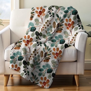 May include: A soft, white throw blanket draped over a white armchair. The blanket features a watercolor pattern of green, brown, and orange leaves and branches. The leaves are in various shades of green, brown, and orange.