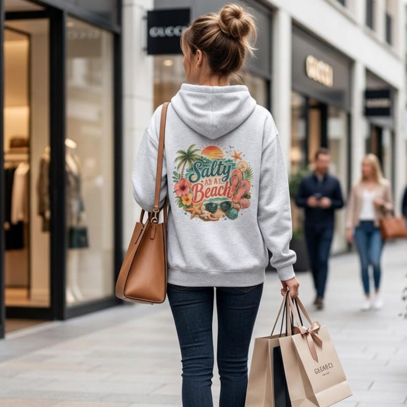 May include: A light gray hooded sweatshirt featuring a circular graphic on the back with the text Salty as a Beach, surrounded by illustrations of palm trees, a sunset, hibiscus flowers, a starfish, flip-flops, and sunglasses.