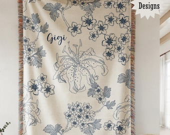 May include: A cream-colored woven throw blanket featuring a dark blue floral pattern and the name Gigi printed in script. The blanket has a fringed edge with multi-colored threads and a star-shaped graphic in the corner with the text Four Designs.