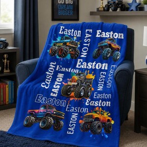 May include: A blue fleece blanket with vibrant monster truck illustrations and the name "Easton" in white. The blanket is draped over a chair. The text "GO BIG DREAM BIGGER" is visible in the background. The blanket has a soft texture.
