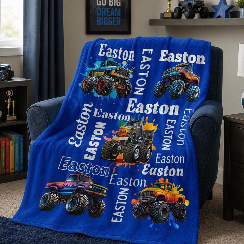 May include: A blue fleece blanket with vibrant monster truck illustrations and the name "Easton" in white. The blanket is draped over a chair. The text "GO BIG DREAM BIGGER" is visible in the background. The blanket has a soft texture.
