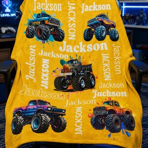 May include: A yellow blanket with monster truck illustrations and the name "Jackson" in white. The blanket displays various monster truck designs with paint splatter effects, and the name is repeated throughout.