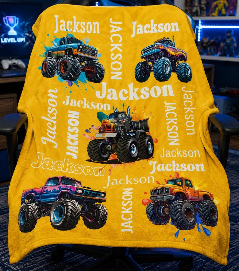 May include: A yellow blanket with monster truck illustrations and the name "Jackson" in white. The blanket displays various monster truck designs with paint splatter effects, and the name is repeated throughout.